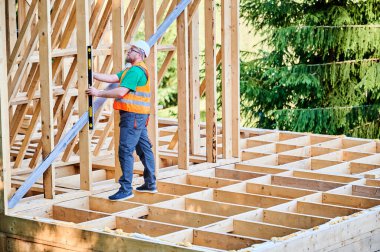 Marangoz iki katlı ahşap çerçeve inşa ediyor. Adam ruh seviyesini kullanarak duvarları kontrol ediyor koruyucu tulum, kask ve turuncu yelek giyiyor. Çevre dostu yapı kavramı.