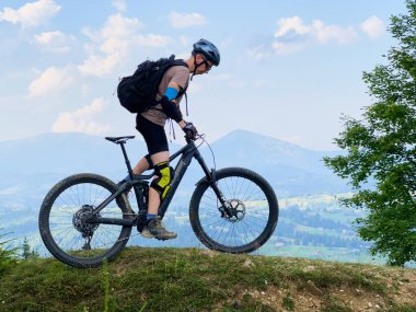 Dışarıda elektrikli dağ bisikleti süren bisikletçi. Erkek turist dağlarda otlak patika boyunca bisiklet sürüyor, başlık ve sırt çantası takıyor. Spor anlayışı, aktif eğlence ve doğa.