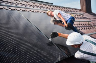İşçiler evin çatısına fotovoltaik güneş paneli inşa ediyorlar. Kasklı ve eldivenli erkek teknisyenlerin güneş modülünü açık alandaki hex anahtarı yardımıyla kurmalarını sağlayın. Yenilenebilir enerji.