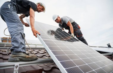 Erkek yükleyiciler fotovoltaik güneş modüllerini evin çatısına kaldırıyorlar. Kasklı elektrikçiler dışarıda güneş paneli sistemi kuruyorlar. Alternatif ve yenilenebilir enerji kavramı.