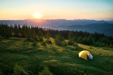 Çimenli yamaçtaki sarı çadırın havadan görünüşü, yükselen güneşle aydınlanıyor. Orman dağlarının nefes kesen manzarası ve berrak gökyüzünün altında yuvarlanan tepeler huzur verici ve pitoresk bir kamp yeri oluşturuyor..