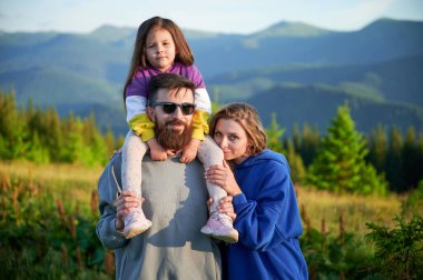 Aile, engebeli dağların arka planında, manzaralı çayırlarda bir arada duruyor. Baba kızını omuzlarında taşır, hem gülümser hem de manzaranın tadını çıkarır, anne ise onların yanında durur..
