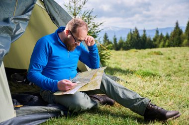 Adam turist çadırlarının girişinde oturuyor, haritaya odaklanmış. Turist pusulayı tutar ve rotasını düşünür. Etrafı ağaçlarla ve bulutlu gökyüzünün altında dağlarla çevrili yemyeşil bir araziyle çevrilidir..