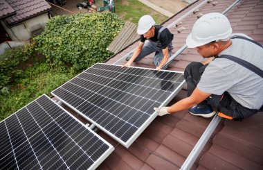 Men technicians mounting photovoltaic solar moduls on roof of house. Engineers in helmets installing solar panel system outdoors. Concept of alternative and renewable energy.
