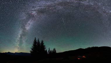Yıldızlar ve Samanyolu ile dolu göz kamaştırıcı gece gökyüzü, sakin kamp alanının üzerinde görkemli bir yay oluşturur. Siluetli ağaçlar ve uzak dağlar manzarayı çerçevelerken, parlayan kamp ateşi sıcak bir dokunuş ekliyor..