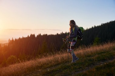 Gün batımında doğa yürüyüşü yapan bir kadın. Dağlarda gezen sportif, pozitif bir kadın. Sırt çantalı, yürüyüş sopası kullanan genç ve sportif bir kadın. Doğayla uyum kavramı.