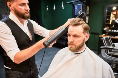 Saç stili için saç kurutma makinesi kullanan deneyimli bir berber. Berber dükkanında düzenli randevusu olan yakışıklı bir adam. Profesyonel kuaför müşterilerinin saçlarını şekillendirirken çekilmiş bir fotoğrafı..