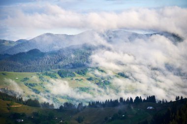 Dağ vadisinin yeşil otlaklar ve dağınık evlerle kaplanmış manzarası. Bulutlar ormanlık tepelerde yavaşça sürüklenir, sakin ve sakin bir atmosfer yaratır..