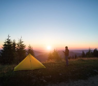 Dağlık kamp alanının üzerinde sakin bir gün doğumu, canlı sarı çadır ve yalnız bir karavan yakınında duruyor. Yumuşak sabah ışığına karşı siluetlenmiş, yuvarlanan tepeler ufka doğru süzülürken,.