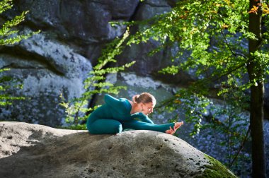 Ormanda yoga yapan bir kadın. Yoga minderinde çıplak ayaklı kadın. Etrafı ağaçlar ve kayalarla çevrili. Bu da meditasyon ya da yoga için ideal barışçıl, doğal bir ortam olduğunu gösteriyor..