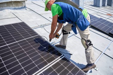 Metal çatı katında fotovoltaik güneş paneli inşa eden bir işçi. Adam teknisyen güneş modülünü açık havada elektrikli matkapla kuruyor. Alternatif enerji üretimi kavramı.