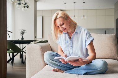 Kanepede rahat bir şekilde oturan bir kadın, kitap okumakla çok meşgul. Arka planında modern estetik olan rahat bir yaşam alanı. Yalnızlığın tadını çıkaran kişi, okumaya dalmış..