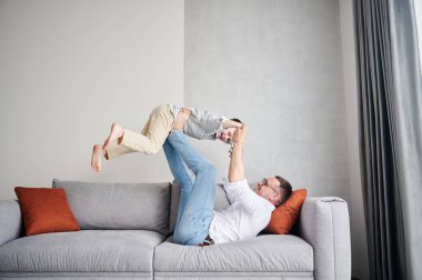 Joyful father lifting happy little son at home. Carefree father and son weekend. Happy young father lying on couch and fooling around with kid.