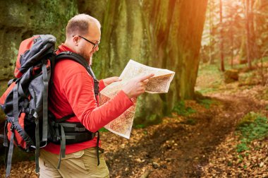Sakallı turist, sık ormandaki büyük kayaların önünde dururken haritayı inceliyor. Gri sırt çantası ve gözlüklü gezgin, açık hava macerası, yürüyüş ya da tırmanma rotası planlıyor..