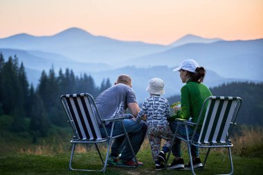 Üç kişilik aile katlanabilir sandalyelerde oturur, dağlarda huzurlu günbatımının tadını çıkarır, doğada birlikte vakit geçirirler. Çocuk ebeveynlerin arasında duruyor. Katmanlı dağ sıraları ve yumuşak, renkli gökyüzü.