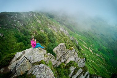 Yürüyüşçü geniş kayaların kenarında, yemyeşil, dik ve kayalık yamaçları olan bir vadide oturuyor. Kadın telefonda görüntü almak için zaman ayırıyor. Etrafı sisle çevrili. Bu olay yerine dinginlik ve manevi kalite katıyor..