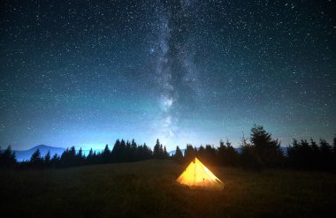 Göz kamaştırıcı yıldızlı gökyüzünün altında çimenli yamaçta parlayan sarı turistik çadır ve Samanyolu belirgin bir şekilde görülüyor. Çam ağaçları ve dağların silueti. Berrak gecenin dingin güzelliği doğada.