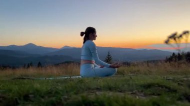 Yoga yapan ve dışarıda, dağlarda meditasyon yapan bir kadın, sakin, doğal bir ortamda. Kadın yoga pozu veriyor paspasın üzerinde, gün doğumunda ya da günbatımında güzel dağ manzarasının arka planıyla.