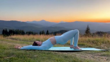 Dağlarda yoga yapan bir kadın, sakin, doğal bir ortamda. Mavi hasırda yoga yapan kadın, gün doğumunda ya da günbatımında güzel dağ manzarasının arkaplanıyla.
