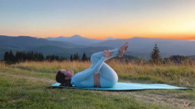 Dağlarda yoga yapan bir kadın, sakin, doğal bir ortamda. Mavi hasırda yoga yapan kadın, gün doğumunda ya da günbatımında güzel dağ manzarasının arkaplanıyla.