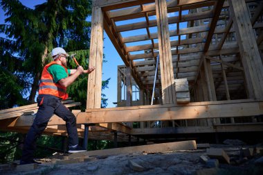 Marangoz ormanın yakınında iki katlı ahşap çerçeve inşa ediyor. Sakallı adam koruyucu kask ve çelik yelek giyerken çivi çakıyor. Modern ekolojik yapı kavramı.
