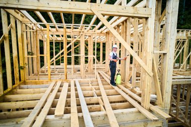 Laborer constructing wooden frame house near forest. Man treating woods, applying fire retardant using sprayer, while dressed in protective suit, helmet. Concept of modern eco-friendly construction. clipart