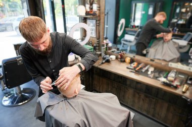 Erkek berber, tarak ve makas kullanarak küçük çocukların saçını kesiyor. Çocuğun saçlarını yetişkin bir erkekten kestiriyor, muhtemelen berber. Modern berber dükkanında profesyonel kuaför ve şirin bir müşteri..