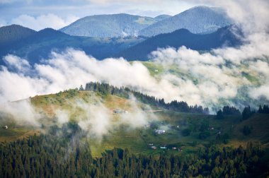 Dağ vadisinin yeşil otlaklar ve dağınık evlerle kaplanmış manzarası. Bulutlar ormanlık tepelerde yavaşça sürüklenir, sakin ve sakin bir atmosfer yaratır..