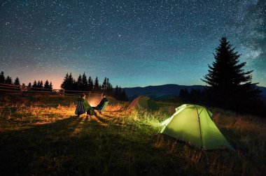 Kamp alanındaki aydınlık çadırların yakınında yıldızlarla kaplı gökyüzünün altında kamp ateşinde dinlenen çift yürüyüşçülerinin silueti. Dağlarda gece kampı, gece gökyüzü büyülü ve huzurlu bir atmosfer yaratır..