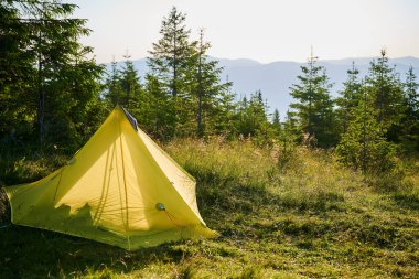 Gün batımının altında çimenli yamaçta turist çadırı. Etrafı genç çam ağaçlarıyla çevrili. Uzak dağların göz kamaştırıcı manzarası ve sabahları huzurlu ve pitoresk bir kamp yeri yaratıyor..