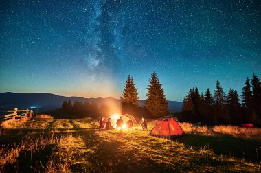 Bir grup arkadaş kamp ateşi etrafında dinleniyor, çarpıcı yıldızlı gökyüzünün altında aydınlık çadırlarla çevrili. Samanyolu uzanıyor ve huzur dolu dağlarda büyülü bir atmosfer yaratıyor..