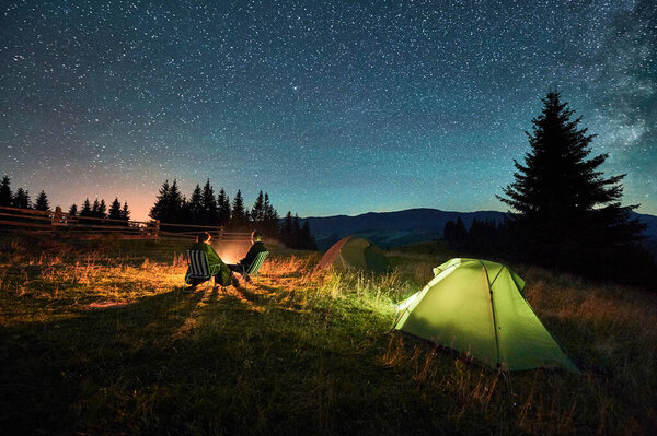 Силуэт пары туристов, отдыхающих у костра под звездным небом, рядом с освещенными палатками в кемпинге. Ночной кемпинг в горах, ночное небо создают волшебную и мирную атмосферу.