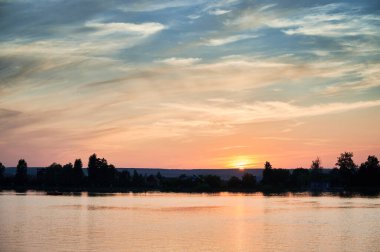 Göz kamaştırıcı günbatımı, pembe, turuncu ve mavi tonlarıyla gökyüzünü aydınlatır. Sakin gölün üzerine sıcak bir ışık saçar. Siluetli ağaçlar ve uzak evler kıyıya vurur, resim gibi ve sakin bir manzara yaratır..