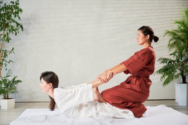 Masör Tayland yogası masajı yapıyor. Geleneksel masaj tedavisi uygulayan kadın terapist, kadın hastaların vücudunu geriyor. Müşteri paspasın üzerinde uzanıyor, sakin atmosferde el masajının keyfini çıkarıyor..