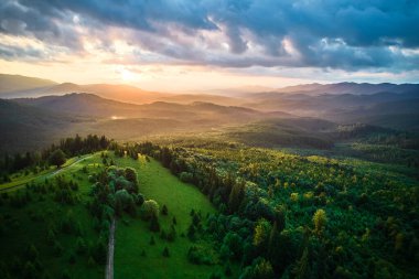 Ormanda hava manzarası nefes kesici gün batımı. Güneş ışınları dramatik bulutları delip geçiyor. Yuvarlanan tepeler ve vadiler boyunca altın parıltılar saçıyor. Uzak dağlar huzur ve resim manzarası yaratıyor..