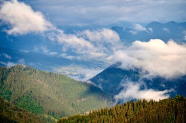 Dağ vadisinin yeşil otlaklar ve dağınık evlerle kaplanmış manzarası. Bulutlar ormanlık tepelerde yavaşça sürüklenir, sakin ve sakin bir atmosfer yaratır..