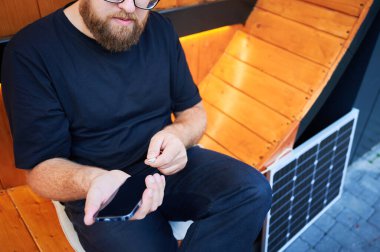 Akıllı telefonu fotovoltaik güneş paneline bağlayan adamın görüntüsü. Sürdürülebilir yenilenebilir enerjinin günlük hayata entegrasyonu, şarj cihazları için güneş enerjisinin pratik kullanımını göstermektedir.
