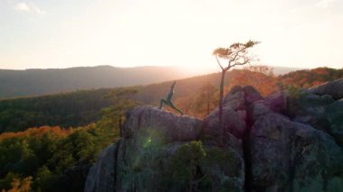 Yoga ve meditasyon yapan bir kadın akşamları dev bir kayanın tepesinde. Güzel günbatımı, sonbahar ormanları, arka planda kayalar ve tepeler. Hava aracı görünümü