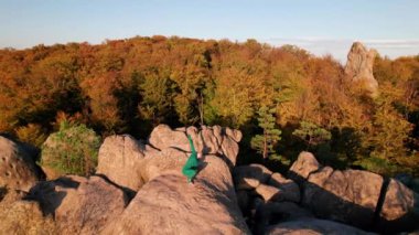 Yoga ve meditasyon yapan bir kadın akşamları dev bir kayanın tepesinde. Güzel günbatımı, sonbahar ormanları, arka planda kayalar ve tepeler. Hava aracı görünümü