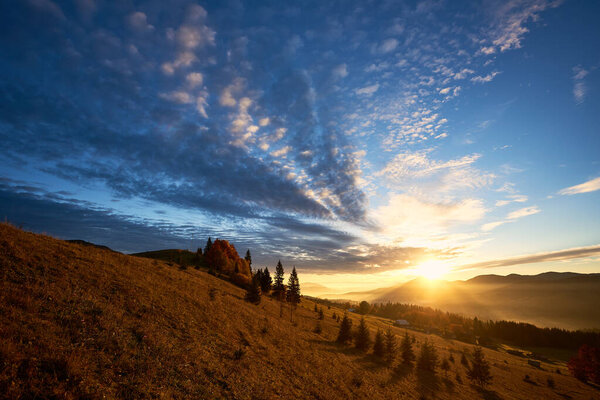 Sun rises over mountains, casting golden rays across landscape. Sky filled with dramatic clouds, creating stunning contrast with clear blue above. Warm light illuminates rolling hills and pine trees.