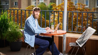 Sakallı adam dışarıdaki yuvarlak masada bir bardak buzlu kahveyle dizüstü bilgisayarda çalışıyor. Yakınlardaki güneş paneli, teknoloji ve sürdürülebilirliği birleştiren sürdürülebilir, modern çevre dostu çalışma alanını vurguluyor.