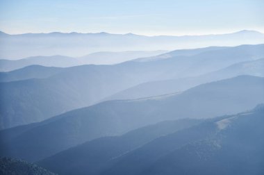 Katmanlı dağ sıralarının yumuşak mavi renkleri berrak gökyüzünün altındaki mesafeye kadar uzanıyor, sakin ve atmosferik bir manzara yaratıyor. Yumuşak eğimler ve uçsuz bucaksızlıkla dolu..