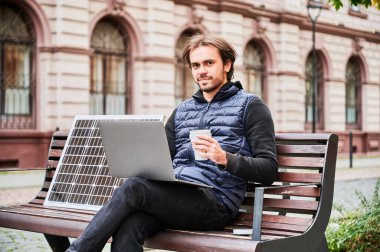 Şehirdeki ahşap bankta oturan adam elinde bir fincan kahveyle dizüstü bilgisayarla çalışıyor. Güneş paneli, tarihi mimarinin arkaplanının ortasında sürdürülebilir, hareketli çalışma alanının vurgulandığı,.