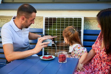 Man shows his family charging smartphone from solar panel. Father, holding phone, points out details on panel. Happy family enjoys moment together. Renewable energy integrate into family life. clipart