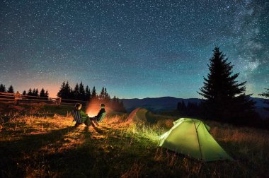Kamp alanındaki aydınlık çadırların yakınında yıldızlarla kaplı gökyüzünün altında kamp ateşinde dinlenen çift yürüyüşçülerinin silueti. Dağlarda gece kampı, gece gökyüzü büyülü ve huzurlu bir atmosfer yaratır..