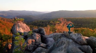 Yoga ve meditasyon yapan bir kadın akşamları dev bir kayanın tepesinde. Güzel günbatımı, sonbahar ormanları, arka planda kayalar ve tepeler. Hava aracı görünümü