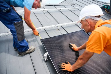 İşçiler fotovoltaik etkilerle elektrik üretmek için evin çatısına güneş paneli inşa ediyorlar. Kurulum için montaj ekipmanlarını ölçmek için cetvel kullanan adamlar.