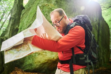 Sakallı turist şaşkın görünüyor. Yoğun ormanda büyük bir kayanın önünde dururken haritayı inceliyor. Gri sırt çantalı ve gözlüklü gezgin, yönünü bulmaya çalışıyor..