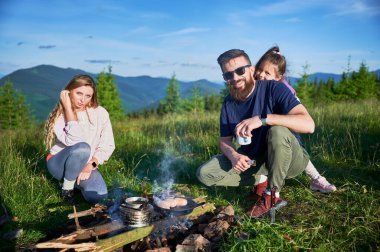 Erkek turist kamp ateşi üzerinde yemek pişirirken kadın ve genç kız resimli dağ çayırında dikiliyorlar. Babam yemekle ilgileniyor. Bereketli yeşillik ve yuvarlanan dağlar berrak mavi gökyüzünün altında.