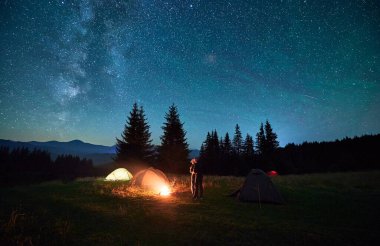 İki yürüyüşçü, arka planda yavaşça parlayan çadırların yanında, yıldızlarla dolu engin gökyüzünün altında kamp ateşinin yanında duruyor. Samanyolu başımızın üstünde uzanıyor, huzurlu dağ ortamında büyülü ve huzurlu bir ortam yaratıyor..